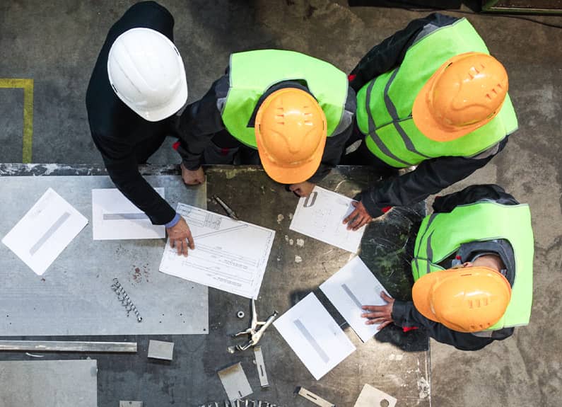 Group of workers on manufacturing floor wearing hard hats and reviewing design drafts at a metal table